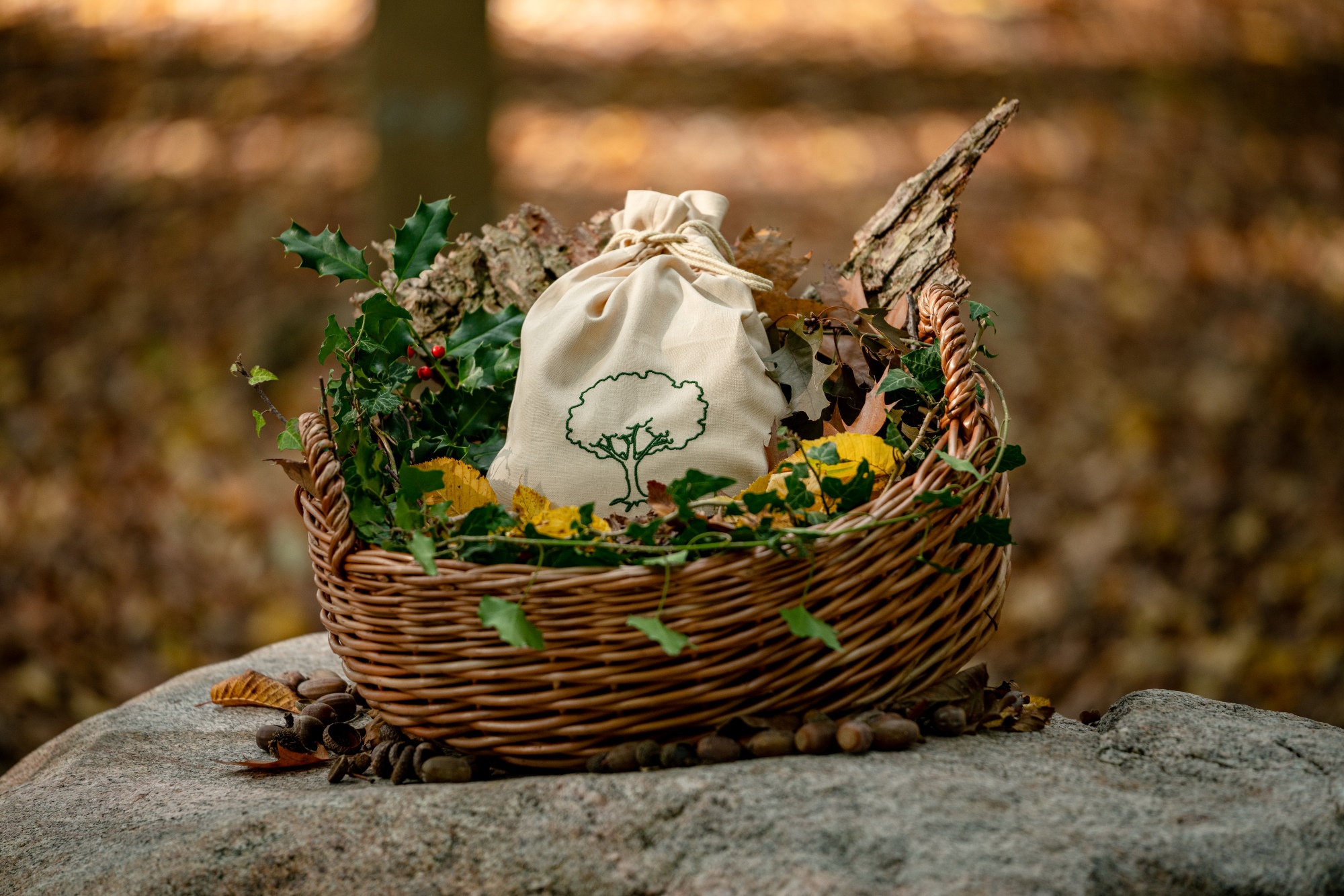 Ruhetuch in einem rustikalem Weidenkorb mit dem Logo vom Ruhewald Sandfort, geschmückt mit Herbstlaub, Stechpalme und natürlichen Elementen auf einem Naturstein-Untergrund.